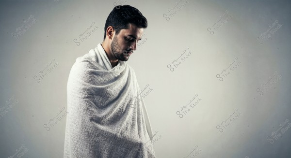 A portrait of a man wearing a white Ihram, standing against a neutral background. The man appears to be in a contemplative state, reflecting a spiritual moment.