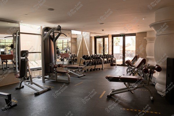 An indoor fitness center with a variety of exercise equipment. The room includes strength training machines, weights, and exercise benches with high ceilings and dark flooring. The walls are equipped with large mirrors and transparent glass windows allowing natural light in. Some outdoor areas are visible in the background through the glass doors.