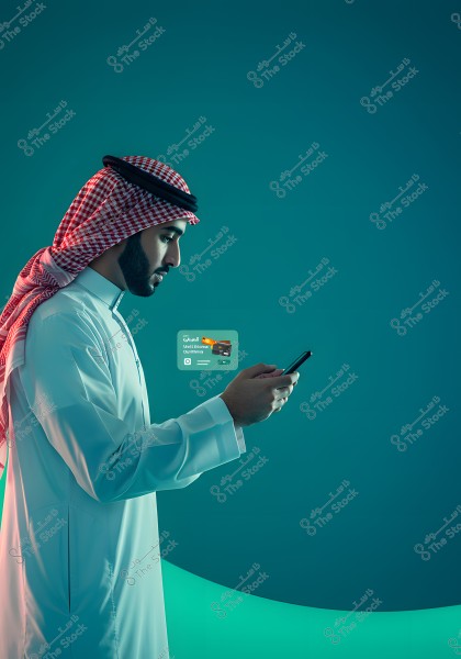Image of a man wearing traditional Saudi Arabian attire, including a white thobe and a red and white checkered keffiyeh. The man is holding and attentively looking at a mobile phone. In the background, a virtual credit card with Arabic text is displayed. The background is blue.