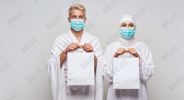 Image of a man and a woman standing side by side, both wearing white Ihram clothing and blue medical masks. The man holds a white bag in his left hand, and the woman holds a white bag in her right hand. The background is simple and plain, emphasizing the two individuals in the foreground.