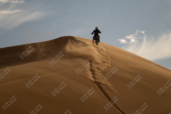 A dirt bike rider traverses a sand dune in the desert. The rider is wearing a helmet and protective clothing, with a blue sky and a few wispy clouds in the background.