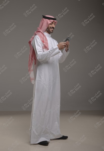 A portrait of a man wearing traditional Saudi attire, including a white thobe, a red checkered shemagh, and a black agal. He is smiling while using his smartphone. The background is a simple gray color.