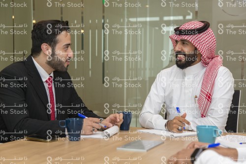 رجل سعودي يرتدي الزي السعودي التقليدي يتحدث مع رجل أجنبي برتدي بدلة رسمية في اجتماع عمل ، رجلان يتناقشان في اجتماع عمل ويدونون الملاحظات