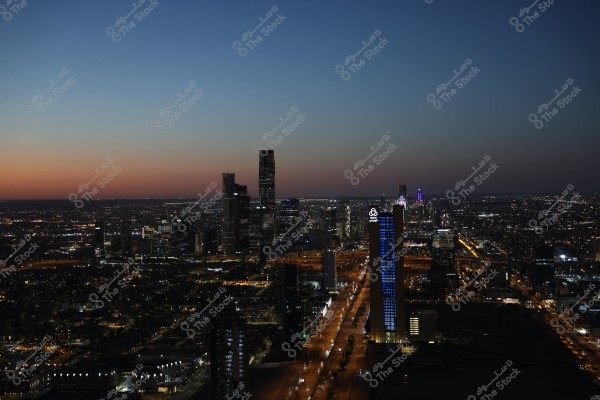 Nighttime image of a large city skyline, showcasing skyscrapers illuminated with lights against a sky transitioning from sunset hues to night. The buildings are lit and streetlights with moving cars can be seen in the distance. A tall tower with a bright screen displaying a logo is prominent in the foreground.