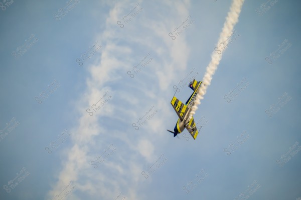 An aerobatic airplane in yellow and blue flies through the sky, leaving a white smoke trail behind. The sky is clear and blue, providing a stunning view of movement and aerial dynamics.