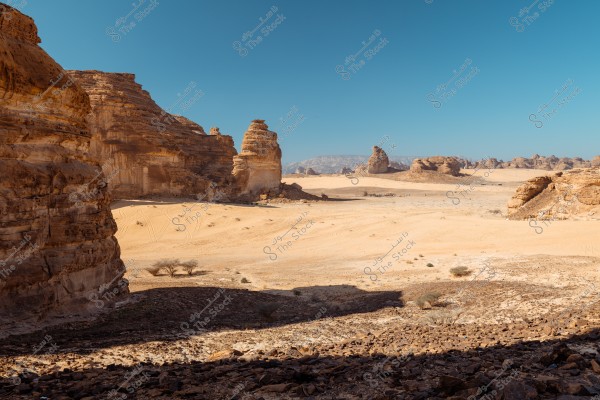 A natural desert landscape featuring massive rock formations in golden brown hues under a clear blue sky. The scene extends with sand, and a few small trees appear in the foreground. The unique and towering rock formations suggest a distinctive natural setting that might be in Al-Ula, Saudi Arabia.