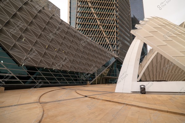 An image depicting a scene of modern architecture featuring unique geometric designs in a broad plaza. The picture includes a structure with reflective glass and interlocking geometric patterns and another building with a slanted white facade, showcasing a modern and futuristic design.