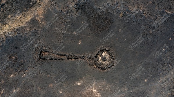 Aerial image of a stone formation in the shape of a key in desert terrain. The ground appears dark brown with the stone formation extending horizontally, with the head of the key on the right side. The surrounding areas are barren with uneven distribution of stones.
