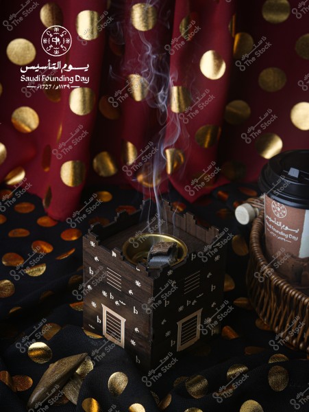 صورة توضح مبخرة خشبية تقليدية ينبعث منها دخان, موضوعة على قماش أسود مزين بالنقاط الذهبية. في الخلفية, يظهر قماش أحمر منقوش بالنقاط الذهبية. إلى الجانب, يوجد كوب قهوة يحمل شعار يوم التأسيس السعودي, وحوله سلة من الخيزران.