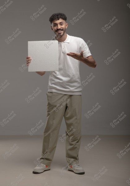 An image of a man standing in a studio with a grey background. He is wearing a white polo shirt, beige trousers, and white sneakers. He holds a square white board in his left hand and points at it with his right hand, smiling at the camera.