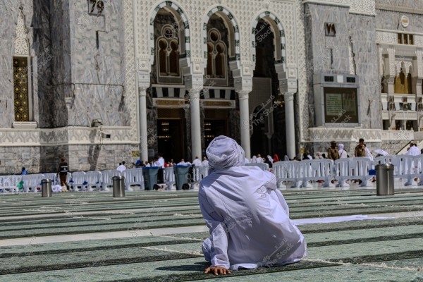 The image shows a person sitting on the ground in the courtyard of the Masjid al-Haram in Mecca. The person is wearing traditional white attire and sits on a ground made of green marble. In the background, part of the mosque\'s facade is visible with its ornate arches and white decorations, along with a group of people walking in the background.