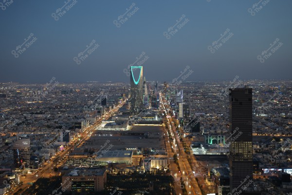 An evening cityscape of Riyadh, Saudi Arabia, featuring the iconic Kingdom Tower illuminated in blue and green hues at the center. Busy streets run along the image with bright city lights, reflecting the vibrant city life.
