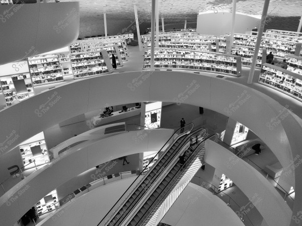An image of a modern library with a contemporary and spacious interior design. The library features tiered and neatly organized bookshelves on multiple levels. An escalator is centrally visible, and natural light floods the area. The lighting accentuates the distinctive ceiling and the dynamic architecture of this cultural space.