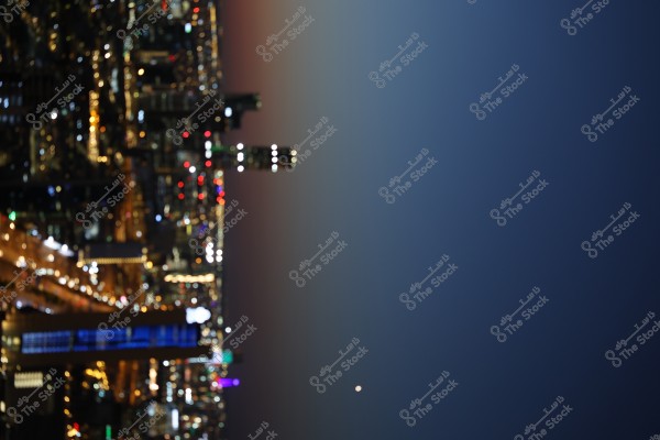 A night view of a city illuminated by the lights of skyscrapers and streets. The sky is a deep blue gradient, and the cityscape appears vibrant with numerous red, white, and golden lights. There is a prominent building with distinct lighting in the center of the city.