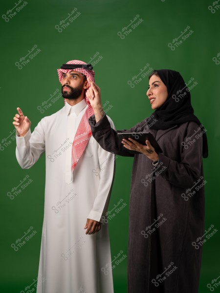رجل وامرأة يرتديان الزي التقليدي السعودي, يقفان أمام خلفية خضراء, المرأة تحمل جهازاً لوحياً.