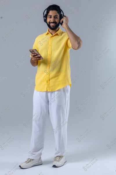 A portrait of a man wearing a yellow shirt and white trousers, listening to music with headphones and holding a smartphone. He stands in a relaxed pose against a white background.