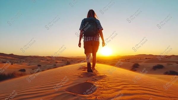 A person walking across sand dunes in the desert during sunset. The person is wearing a checkered shirt, shorts, and carrying a backpack. The image showcases a breathtaking view of the expansive golden sands, wide horizon, and light blue sky.