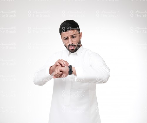 بورتريه رجل سعودي يرتدي الزي السعودي التقليدي يضع النظارات و ينظر للساعة بينما يمسك شاشتها بيده متعجبًا