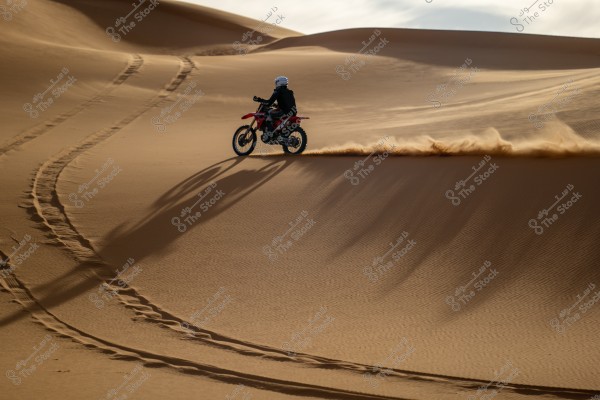 A motorcycle riding across sandy desert dunes, leaving a trail of dust behind in the soft sand under the sunlight. The sky in the background is partially clear.