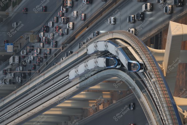 Aerial view of a busy road filled with numerous vehicles and several lanes. A modern metro train is visible traveling on a track above the highway. The architectural design of the roads and bridges appears complex and synchronized, with the evening glow casting gentle shadows on the road. The image seems to depict a modern city with advanced urban planning.
