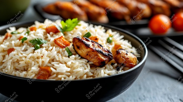 An image of a bowl of rice garnished with pieces of grilled chicken and colorful vegetables like carrots and green cilantro. The dish is served in a dark-colored bowl placed on a grey wooden table, with a blurred background featuring tomatoes and chicken pieces.