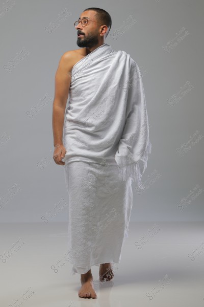 A portrait of a man wearing the white Ihram clothing, which consists of two pieces of unstitched white fabric. The man stands barefoot on a smooth white floor and gazes to his side. He is wearing glasses and has a short beard. The background is gray and neutral. The image reflects the ambiance of preparing for Hajj or Umrah in Saudi Arabia.