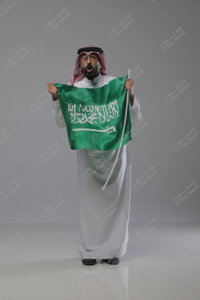 A portrait of a man wearing traditional Saudi attire, featuring a white thobe, a red-and-white checkered keffiyeh, and a black agal. He is holding the Saudi Arabian flag enthusiastically, against a light gray background.