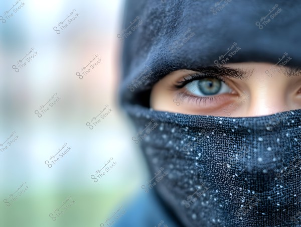 A close-up image of a woman\'s eye wearing a black hijab covering the hair and part of the face, with a fabric patterned with small white speckles covering the lower part of her face. The background is blurred, highlighting the focus on the blue eye.