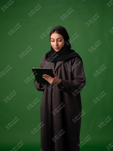 Portrait of a woman wearing a black abaya and a black hijab, standing while holding a tablet. The background is green. Her appearance suggests she may be from a Gulf country.