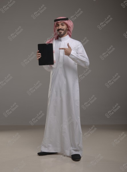 A portrait of a man wearing a Saudi thobe and a red and white ghutra, smiling in a studio setting. He is holding a tablet and pointing at it with one hand. The background is plain gray.