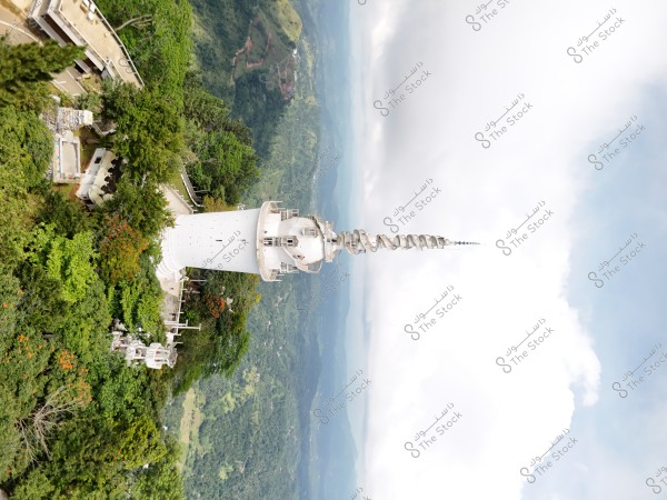 An image of a tall white tower located in a green mountainous area, surrounded by trees and vegetation. The tower extends vertically with a distinctive spiral top. In the background, there is a natural view of wide green landscapes and a blue sky filled with clouds.