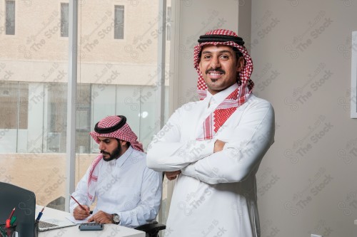 رجل سعودي يرتدي الزي السعودي التقليدي يقف مكتوف اليدين أثناء إجتماع في العمل نهارًا