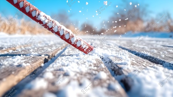 A close-up view of a red stick decorated with snow being used to clear snow from a wooden surface. The image shows snowflakes scattered in the air with a blurred background of a blue sky and a snow-covered ground.