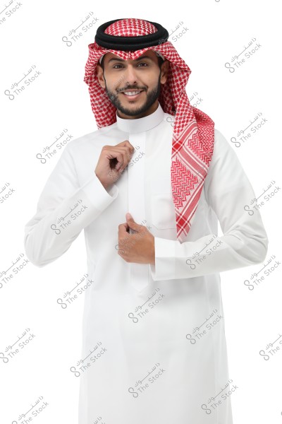 Image of a man wearing traditional Saudi attire, including a white thobe, a red and white checkered shemagh, and a black agal. The man is smiling and looking at the camera, placing his hand on the front of his thobe as he adjusts it. The background is plain white.
