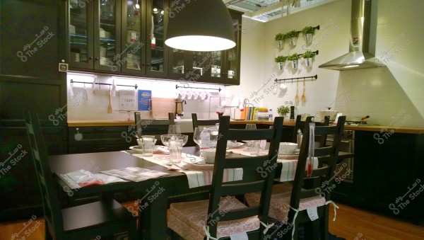 A modern and stylish kitchen featuring a black wooden dining table surrounded by four chairs. The table is neatly set with glassware, dishes, and cutlery. In the background, there are dark-colored cabinets and hanging kitchen utensils alongside small potted plants on the wall. There is also a stainless steel range hood above a gas stove. The space is illuminated by a large hanging lamp in the center.