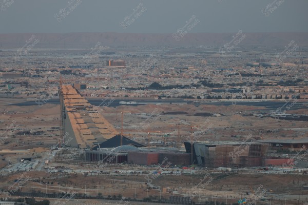 منظر جوي لمدينة كبيرة في الصحراء, يحتوي على مبنى معماري كبير يشبه المثلث وقيد الإنشاء. المبنى محاط بالعديد من الرافعات والطرق. في الخلفية, تظهر منازل ومباني المدينة, وأفق جبلي بعيد تحت سماء غائمة.