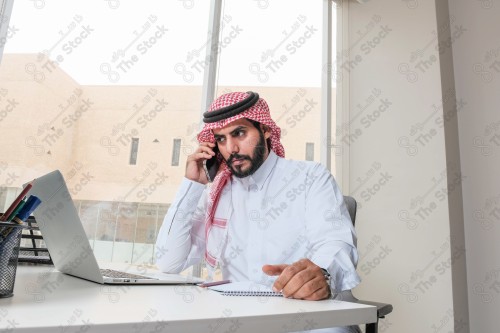 رجل سعودي يرتدي الزي السعودي التقليدي يعمل في مكتب بواجهة زجاجية ويستخدم قلم رصاص لتدوين الملاحظات