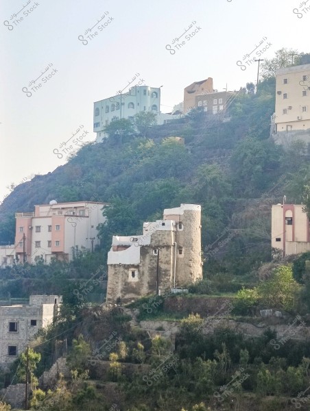 An image depicting a cluster of traditional and modern houses situated on a hillside covered with green vegetation. The buildings vary in style and color, with some featuring white, light blue, and beige hues. Electrical wires stretch across the hill. The surrounding hillsides are covered with forest and greenery.