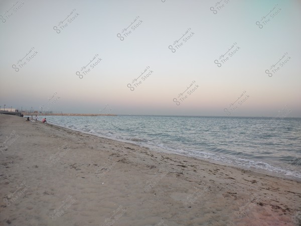 A sandy beach stretches into the calm sea horizon under an open sky with soft twilight colors. A few individuals are sitting on the beach near the water, while some lamps are visible in the background on the left, with a clear breakwater extending from the beach into the sea.