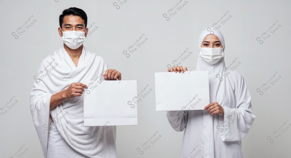 The image shows a man and a woman wearing traditional white clothing for pilgrimage, with the man in an Ihram and the woman in a white veil. Both are wearing white face masks. The man holds a white paper bag in his right hand, while the woman holds a similar white paper bag. The background is a plain white, emphasizing the couple. The attire suggests Islamic culture and may be associated with Saudi Arabia or other Islamic countries.
