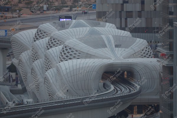 An image of a train station with a modern and sophisticated architectural design, featuring a wavy roof with mesh ventilation panels. The railway track enters through an opening in the building. Roads and modern buildings can be seen in the background.