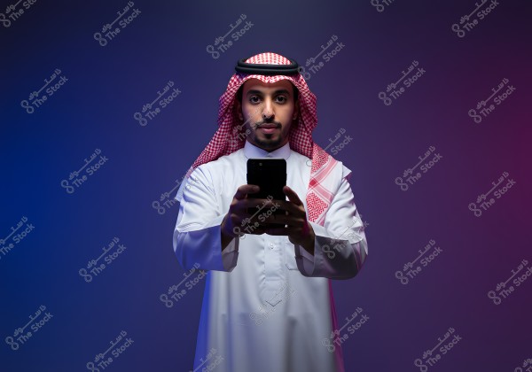 An image of a man wearing a white thobe, a red checkered shemagh, and a black agal, holding a mobile phone in his hands against a colorful background with shades of blue and purple. Bright lighting highlights the details of the traditional attire.
