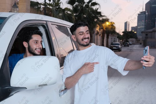 A snapshot of two young Saudis chatting and having fun on the road, their features showing joy and happiness, taking a selfie with a mobile phone