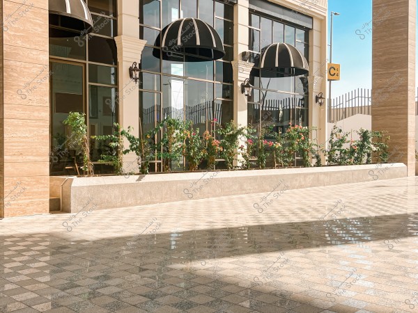 A building with a large glass facade, adorned with black awnings. In front of the building is a small garden with a variety of flowering green plants. The ground is polished and glistens under the sunlight. There is a pillar visible on the left side and a sign that reads \"C4\" attached to the building.