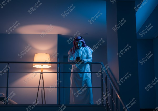 Image of a man standing by an indoor balcony of a modern design at night. The man is wearing traditional Arab attire, including a thobe and ghutra, and is focused on a mobile phone in his hand. The space is dimly lit by a floor lamp, creating a serene atmosphere in the room.