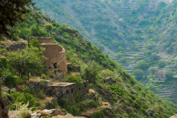 A scenic view of hills covered in dense green vegetation with traditional mud and stone buildings on the hillside. Agricultural terraces are visible in the background, highlighting the beauty of rural nature and traditional architecture in a mountainous region.