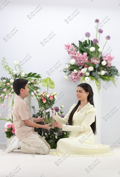 An image shows a woman in a light-colored dress sitting on the ground, receiving a bouquet of flowers from a young boy wearing a short-sleeved shirt and light-colored pants. The background is adorned with a variety of colorful flowers, including pink and white roses, orchids, and anthurium.