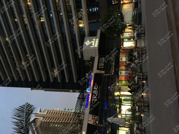 The image shows a modern commercial complex in the evening, with its facade illuminated by bright lights. There is an outdoor café with several tables and chairs arranged neatly. The signs on the building are in both Arabic and English. Palm trees line the front, giving the place a Gulf region ambiance.
