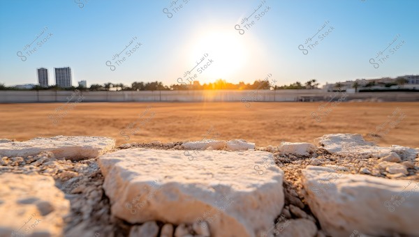 صورة لغروب الشمس فوق منظر طبيعي صحراوي, مع مقدمة للصورة تظهر صخور وحصى قريبة. في الخلفية, يظهر جدار منخفض وأبنية بعيدة مع أشجار بالأفق. السماء زرقاء صافية مع وهج الشمس الساطعة.