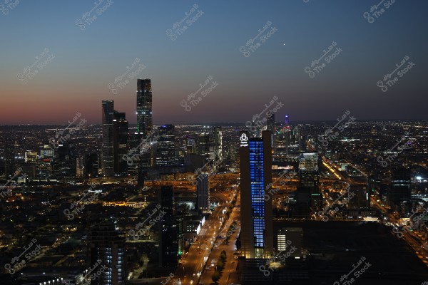 A night view of Riyadh\'s skyline, featuring illuminated skyscrapers under the city lights. Prominently visible is a tall building lit in blue bearing the \"Al Rajhi\" logo. Wide streets stretch between the buildings, with bright lights shining in the evening skyline under a dark sky with remnants of sunset light on the horizon.